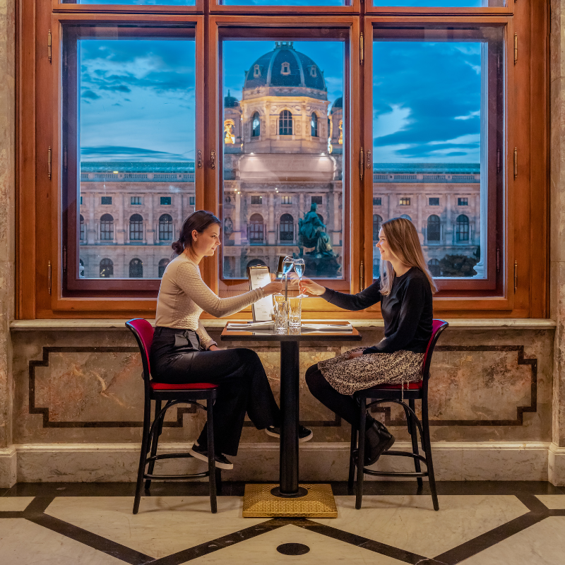 Zwei Frauen die bei Sonnenuntergang an einem Fensterplatz sitzen und mit Sekt anstoßen