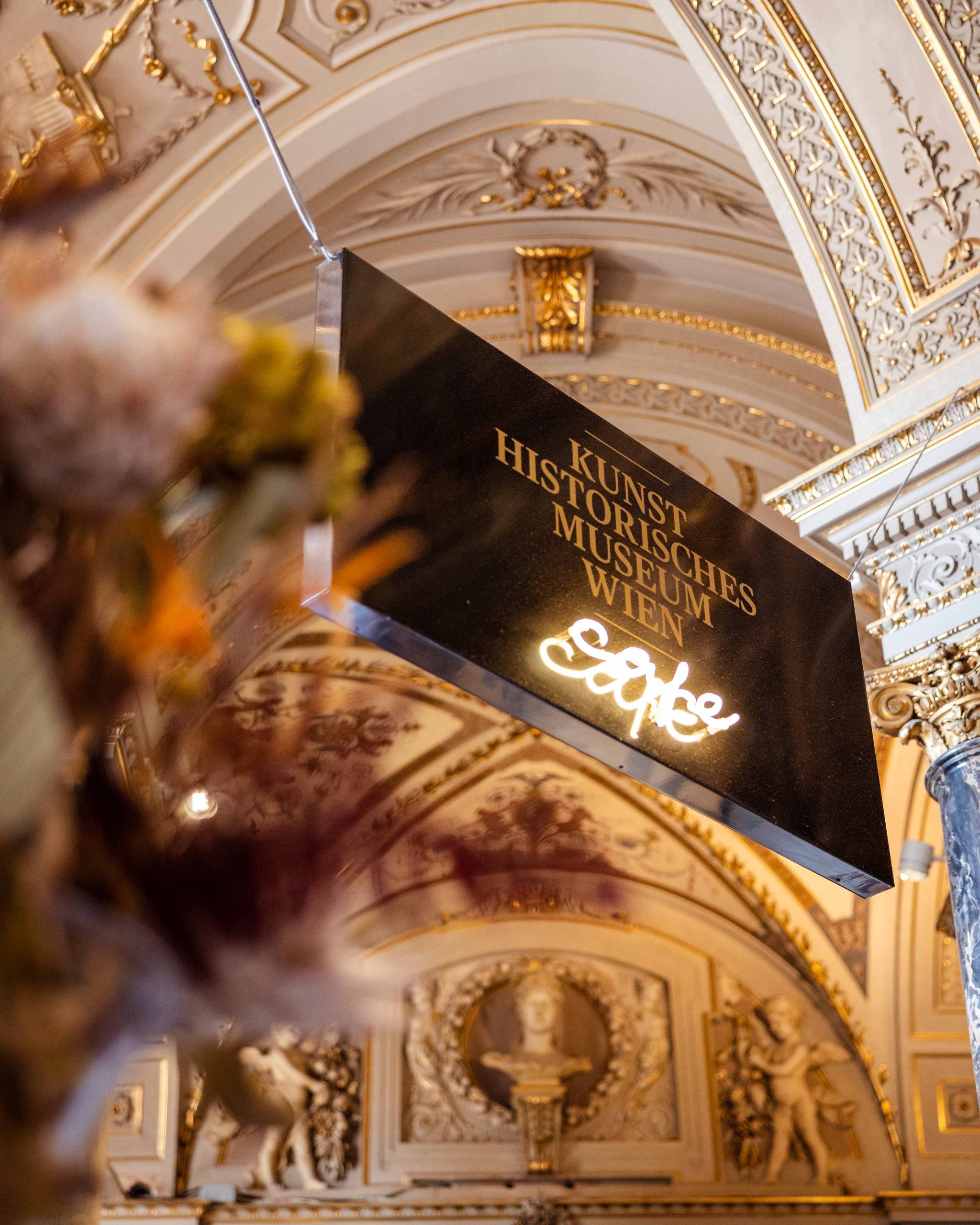 Beleuchtetes Schild das an einer Stuckdecke hängt, Aufschrift Kunsthistorisches Museum Wien Café